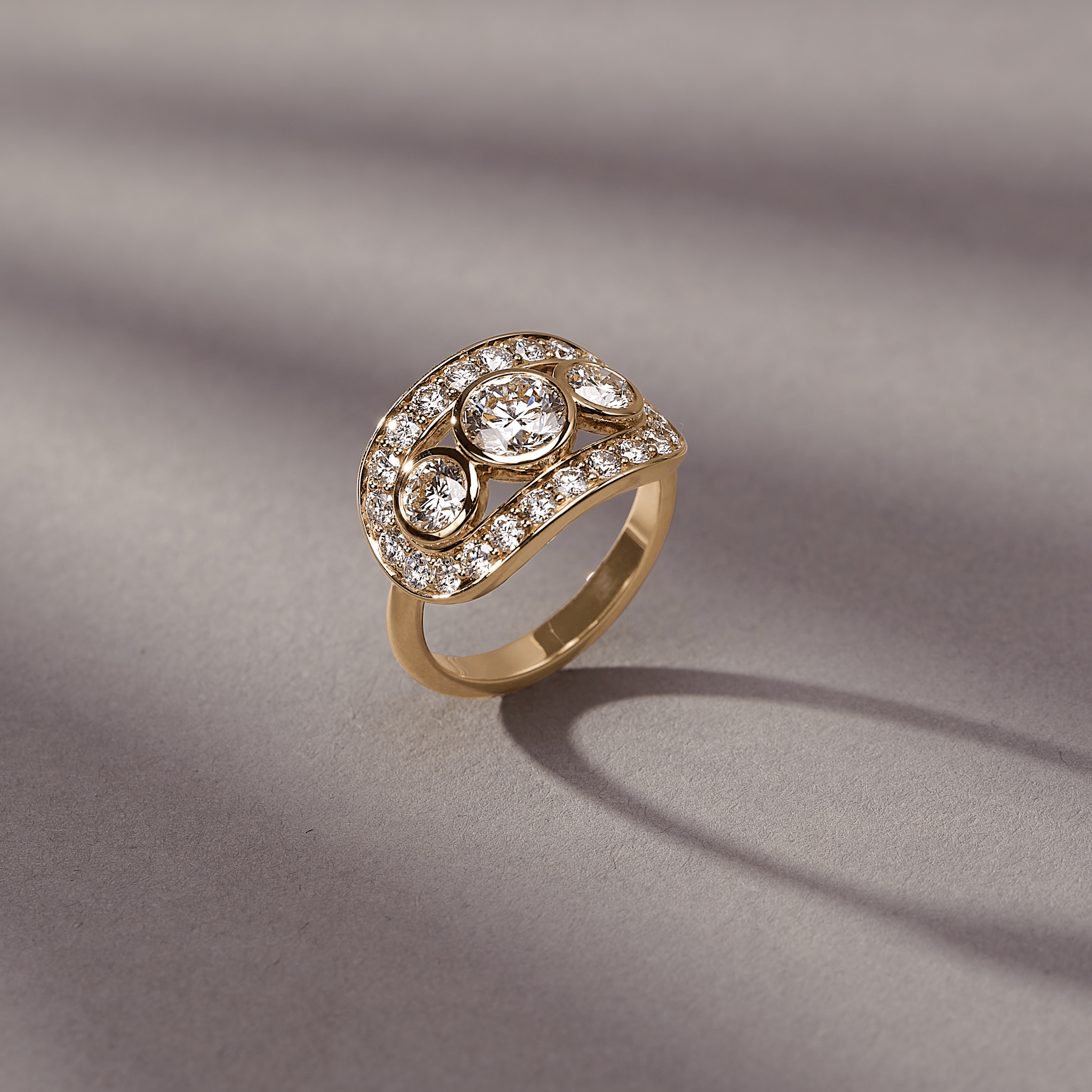 Heritage Trio Ring i 18kt guld fotograferet i vinkel på en struktureret grå baggrund med bløde skygger.