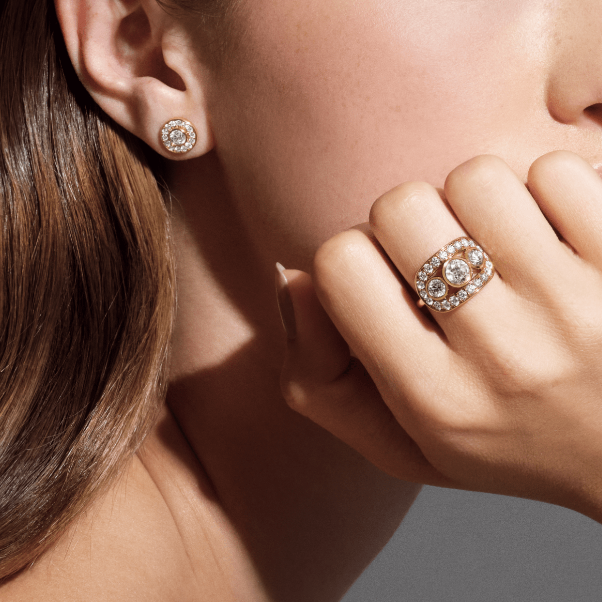 Heritage Trio Ring båret på hånden holdt op mod øret, med en diamantbesat stud-ørering synlig i baggrunden.