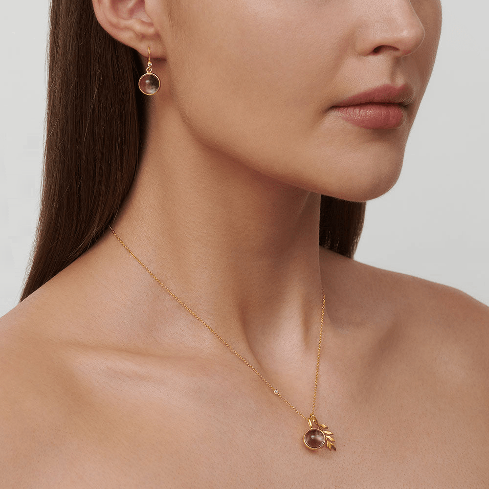 Model bærer Prime øreringe i guld med rosa cabochonslebet krystal, kombineret med matchende vedhæng i halskæde