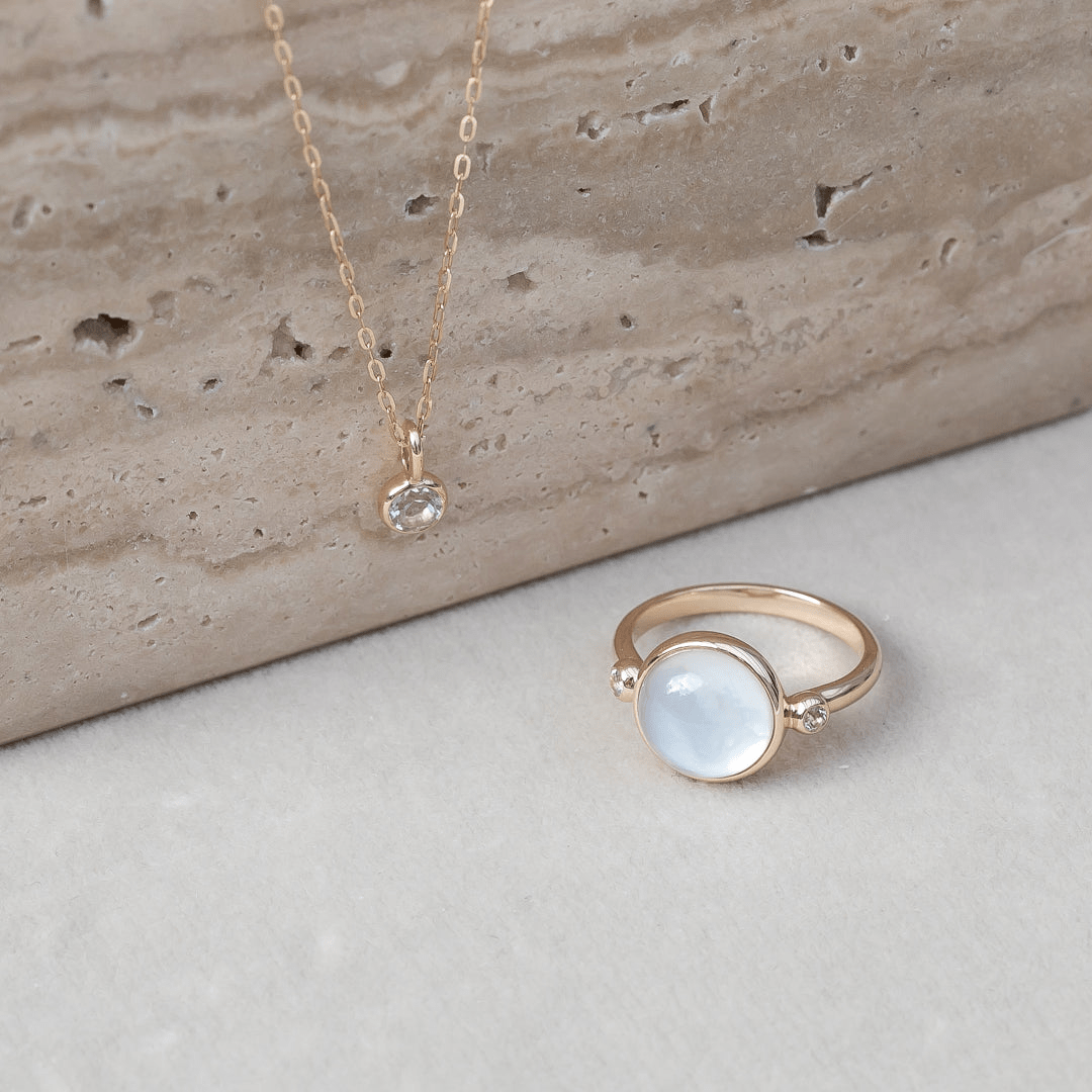Carla Ring i guld med hvid cabochon-slebet sten, foto ved siden af en guldkæde med vedhæng