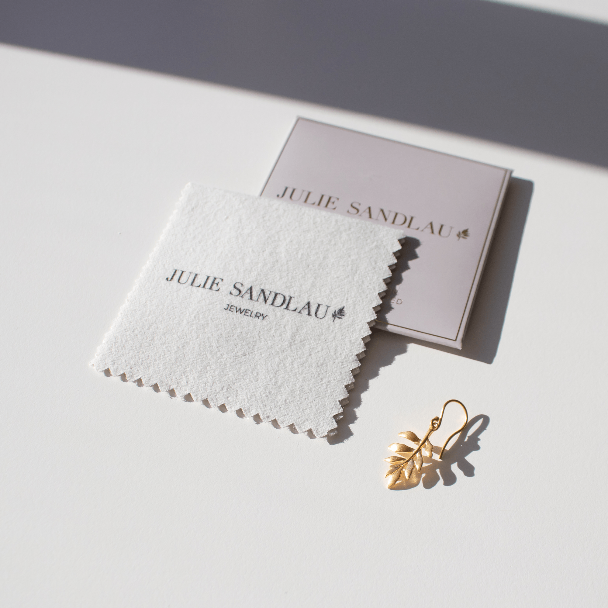 Julie Sandlau poleringsklud med logo ved siden af mærkekort og et lille bladformet smykke