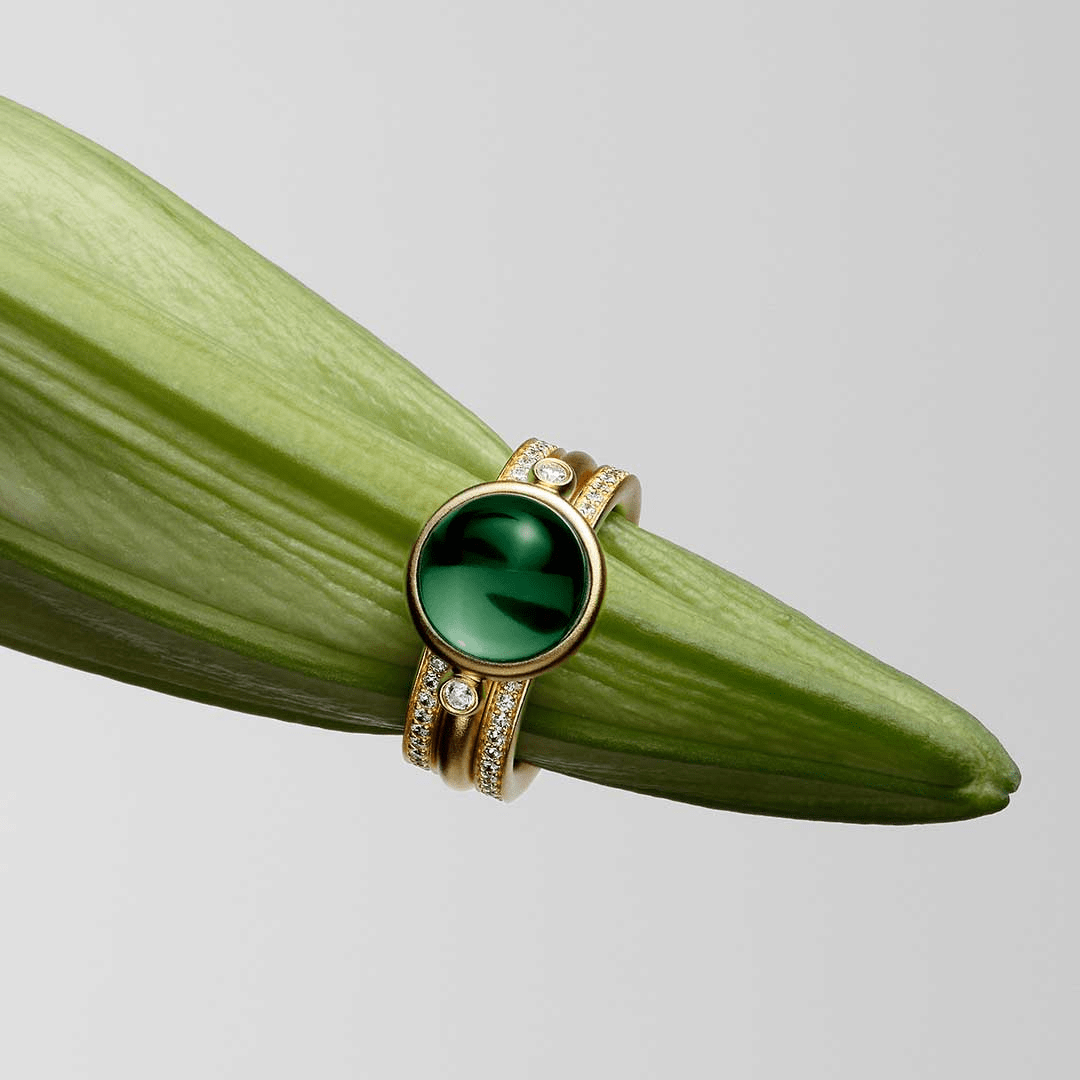 Prime double Infinity Look-ringsæt holdt op mod et grønt blad, grøn sten i guldfatning med diamantbesatte bånd